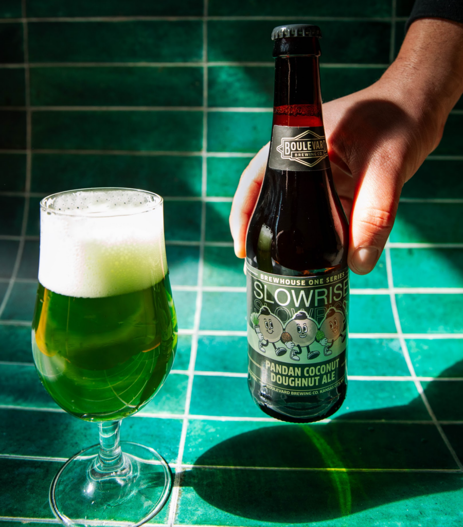A hand places a bottle of Boulevar Slowride Brewhouse One Series Pandan Coconut Doughnut Ale on a green tiled surface. Next to it, a glass of green beer with a frothy top sits on the table, catching sunlight.